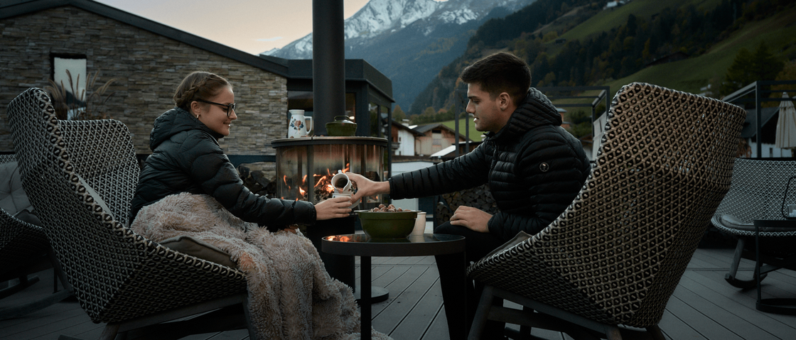 Sich erholen im Hotel in Neustift im Stubaital Paar sitzt draußen vor Feuerstelle in den Bergen, umgeben von Stühlen und Decke