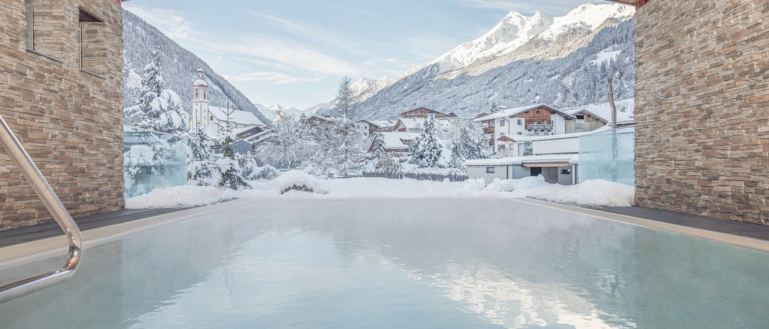 Entspannen im Wellnesshotel im Stubaital Blick auf winterliches Bergdorf von einem beheizten Außenpool mit Steinmauern